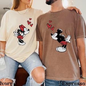 May include: Cream and brown t-shirts featuring Mickey and Minnie Mouse kissing, with red heart graphics. The shirts have the words "Ivory" and "Espresso" printed at the bottom. The shirts are a relaxed fit.