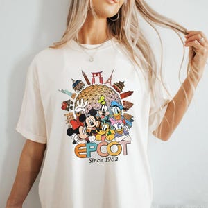 Pu&ograve; includere: T-shirt color crema con una grafica colorata dei personaggi Disney, tra cui Topolino e Minnie, Pippo, Paperino e Paperina. La grafica include la parola "EPCOT" e il testo "Since 1982" con immagini di monumenti.