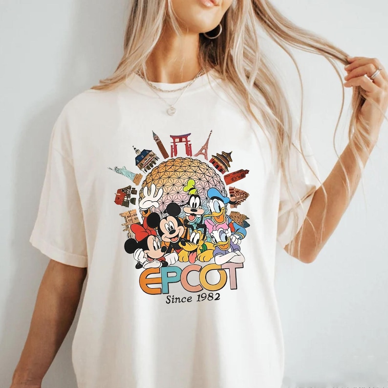 Pu&ograve; includere: T-shirt color crema con una grafica colorata dei personaggi Disney, tra cui Topolino e Minnie, Pippo, Paperino e Paperina. La grafica include la parola "EPCOT" e il testo "Since 1982" con immagini di monumenti.