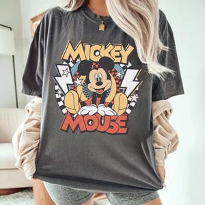 May include: Dark gray t-shirt featuring a colorful graphic of Mickey Mouse. The design includes the words "Mickey Mouse" in yellow and red, with Mickey in a seated pose. The shirt has a vintage, worn look.