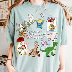 May include: A light green t-shirt featuring a graphic print of various Toy Story characters including Buzz Lightyear, Woody, Jessie, Rex, Slinky Dog, Hamm, Bullseye, and an Alien. The text "YOU'VE GOT A FRIEND IN ME" is displayed in the center.