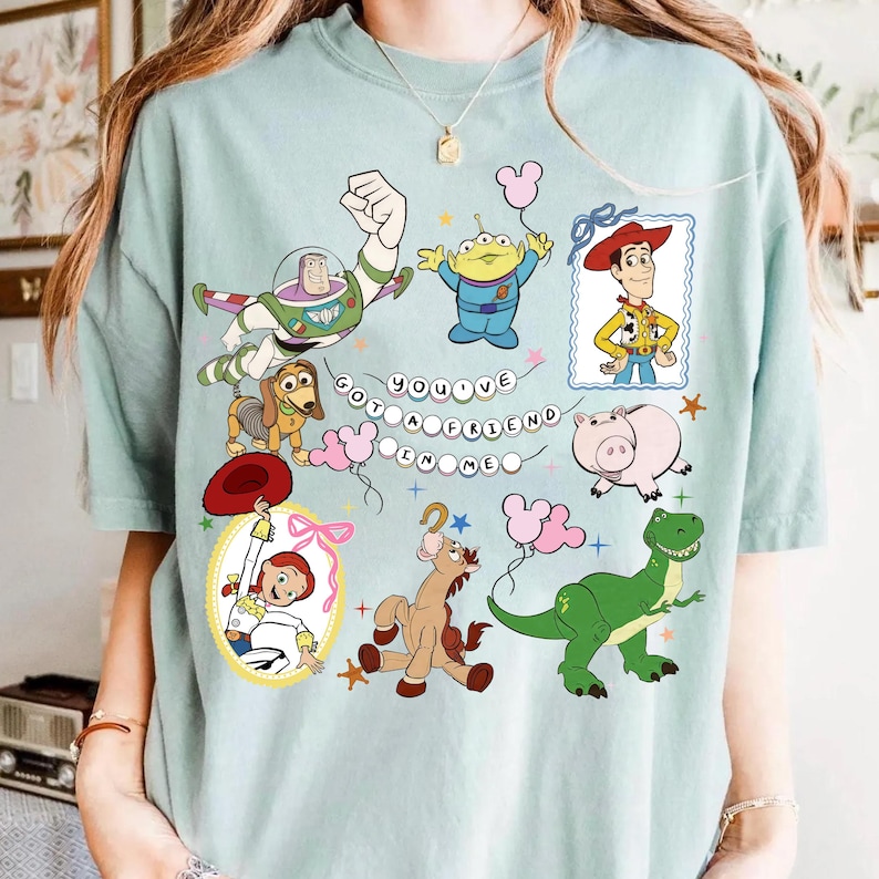 May include: A light green t-shirt featuring a graphic print of various Toy Story characters including Buzz Lightyear, Woody, Jessie, Rex, Slinky Dog, Hamm, Bullseye, and an Alien. The text "YOU'VE GOT A FRIEND IN ME" is displayed in the center.