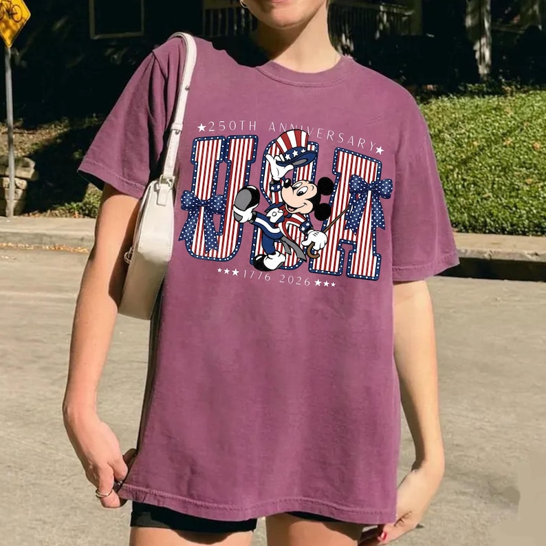 May include: Maroon t-shirt with a graphic of Mickey Mouse in patriotic clothing, holding a golf club. The word "USA" is in red and blue stripes, with "250th Anniversary" and "1776 2026" text.