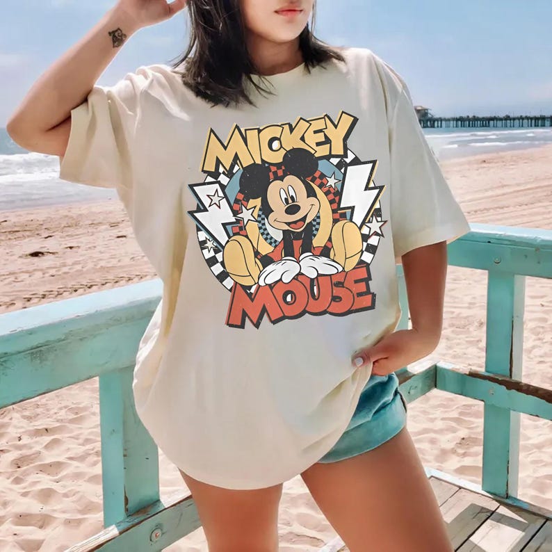 May include: Cream-colored t-shirt featuring a graphic of Mickey Mouse with the text "Mickey Mouse". The design includes a checkered background, lightning bolts, and stars. The shirt is worn on a beach.