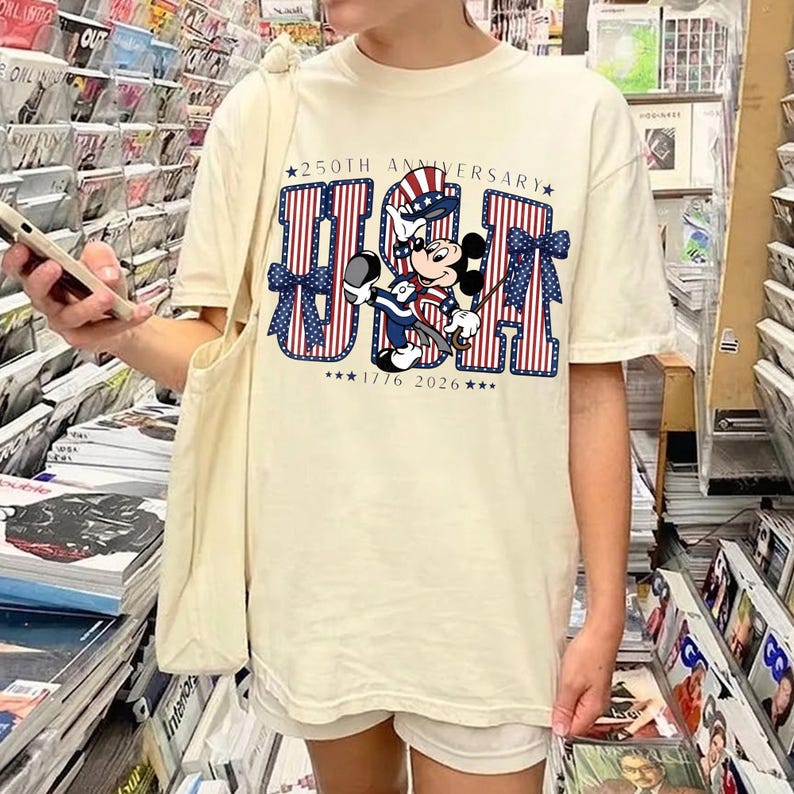May include: A cream-coloured t-shirt with a patriotic design. The design features the letters "USA" in red and blue stripes, with blue bows and stars. A cartoon character in patriotic clothing is included, celebrating the 250th anniversary.