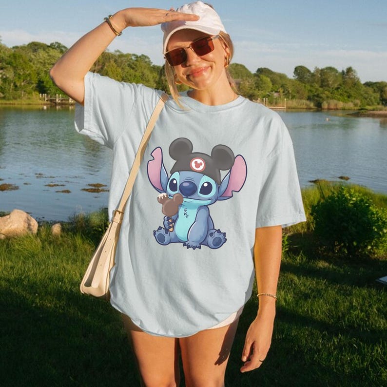 May include: Light blue t-shirt featuring a cartoon character, Stitch, wearing a black hat with a red emblem and holding a brown ice cream cone. The character is blue with large ears. The shirt is worn by a person outdoors.