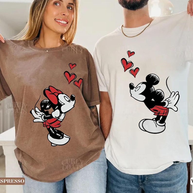 May include: Two t-shirts with cartoon character designs. One is brown with Minnie Mouse, red hearts, and a red bow. The other is white with Mickey Mouse and red hearts. The shirts are a matching set.