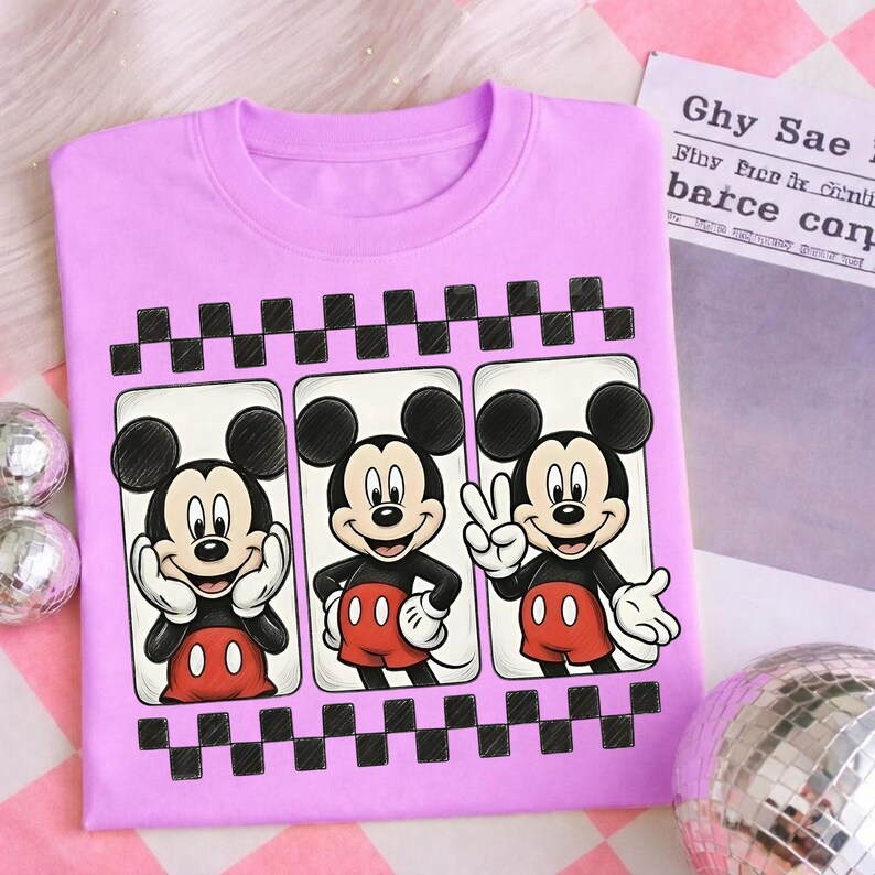 May include: A light purple sweatshirt featuring three panels with Mickey Mouse illustrations. Each panel showcases a different pose of Mickey, with a black and white checkered pattern above and below the panels. The sweatshirt is a casual wear item.