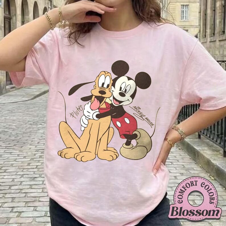 May include: Light pink t-shirt featuring a graphic of Mickey Mouse hugging Pluto. Mickey Mouse is wearing red shorts and Pluto is tan. The text "Mickey Mouse" and "Pluto" are also visible on the shirt. The shirt has the text "Comfort Colors Blossom" on the bottom right.