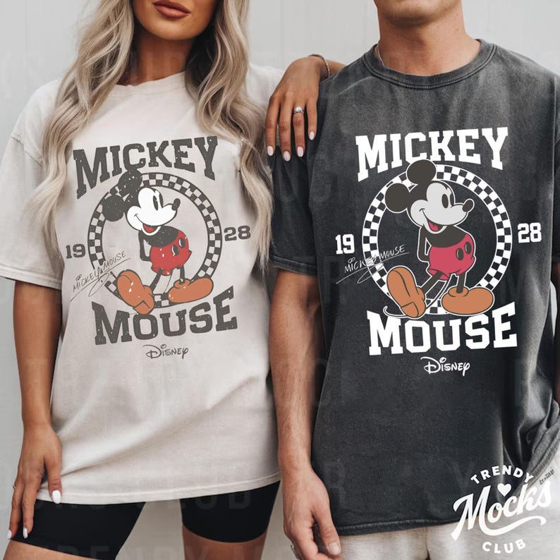 Peut inclure: Deux t-shirts avec un motif Mickey Mouse. Le design comprend le texte "MICKEY MOUSE" dans un cadre circulaire avec les ann&eacute;es "19" et "28". Le logo Disney est en bas. Un t-shirt est beige, l'autre est gris fonc&eacute;.
