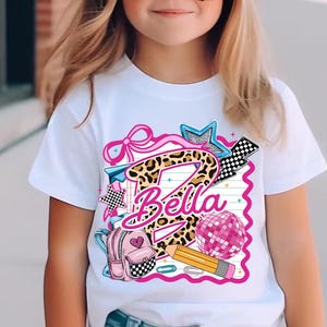 Pode incluir: Uma t-shirt branca de manga curta com um design gráfico que apresenta uma letra B grande com estampado de leopardo, o nome Bella em letra cursiva rosa, uma mochila rosa, um lápis amarelo, uma bola de discoteca rosa, um laço rosa e xadrez.