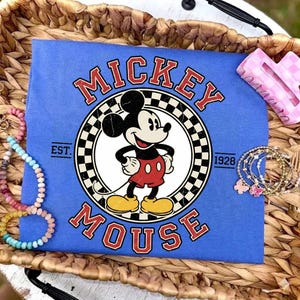May include: A blue t-shirt with a Mickey Mouse graphic. The design features the words "Mickey Mouse" in red, a checkered circle, and the text "Est. 1928". The shirt is displayed with accessories.