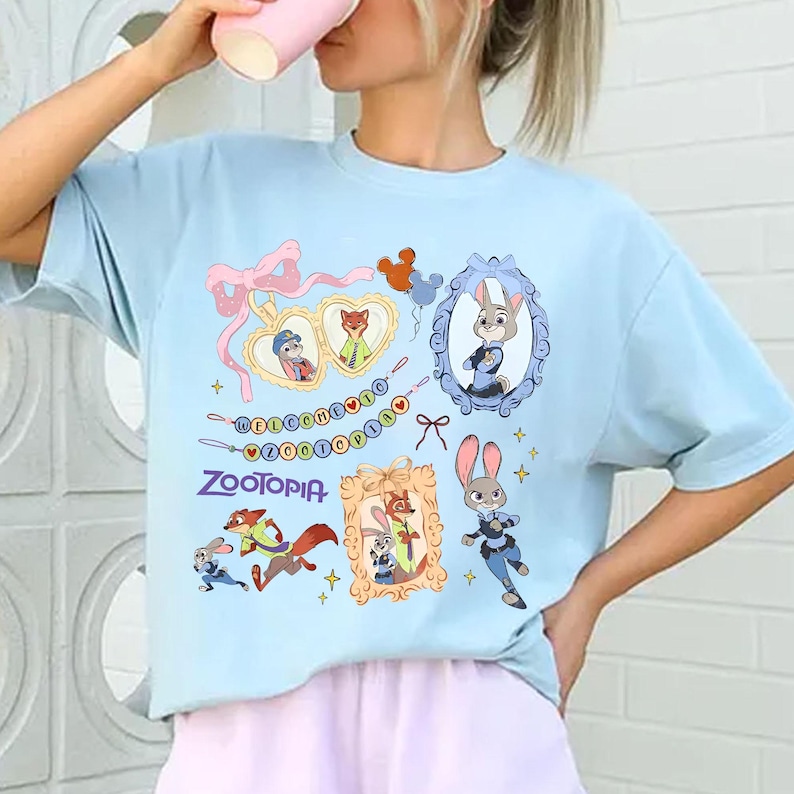 May include: Light blue t-shirt featuring Zootopia characters and the word "Zootopia". The design includes framed images of Judy Hopps and Nick Wilde, along with other characters and decorative elements.