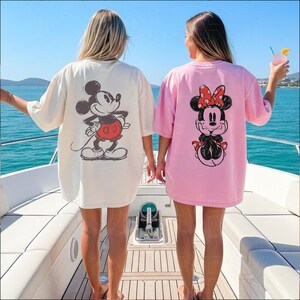 May include: Two oversized t-shirts, one cream and one pink, each featuring a Disney character graphic on the back. The cream shirt has Mickey Mouse, and the pink shirt has Minnie Mouse. The shirts are worn on a boat with a blue ocean background.