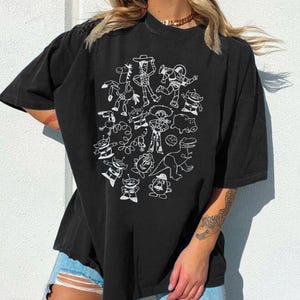 May include: Black t-shirt featuring a white outline illustration of characters from a popular animated movie. The design includes a cowboy, space ranger, aliens, and other recognizable figures. The shirt has short sleeves and a relaxed fit.