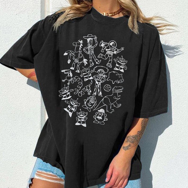 May include: Black t-shirt featuring a white outline illustration of characters from a popular animated movie. The design includes a cowboy, space ranger, aliens, and other recognizable figures. The shirt has short sleeves and a relaxed fit.