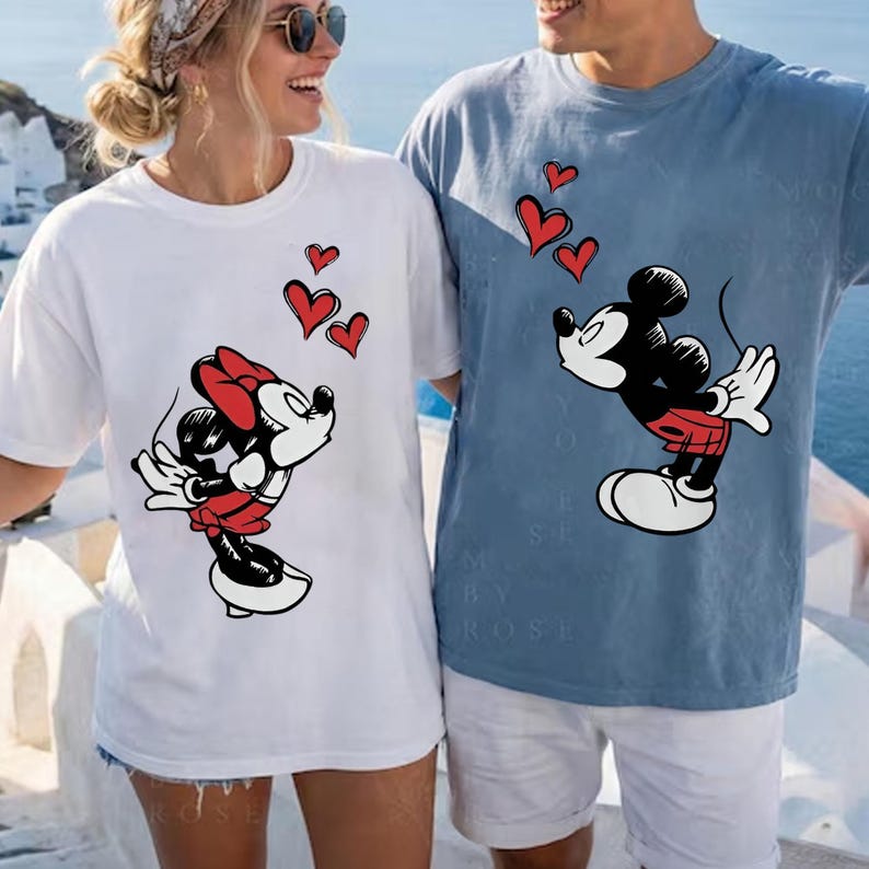 May include: White and blue t-shirts featuring Mickey and Minnie Mouse designs. The Mickey Mouse design is on a blue shirt, and the Minnie Mouse design is on a white shirt. Both designs include red hearts.