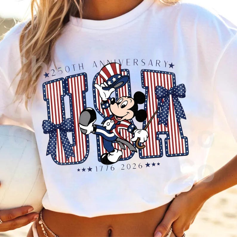 May include: White t-shirt with a patriotic "USA" design in red and blue stripes, adorned with blue bows and stars. A cartoon mouse character, dressed in patriotic clothing, is holding a cane and a hat. The shirt celebrates the 250th anniversary.