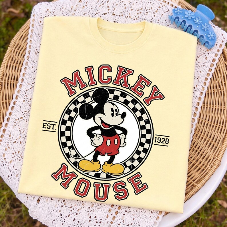 May include: A pale yellow t-shirt with a Mickey Mouse graphic. The design includes the words "Mickey Mouse" in red, a black and white chequered circle, and the year "1928". A blue hair clip is visible in the top right corner.