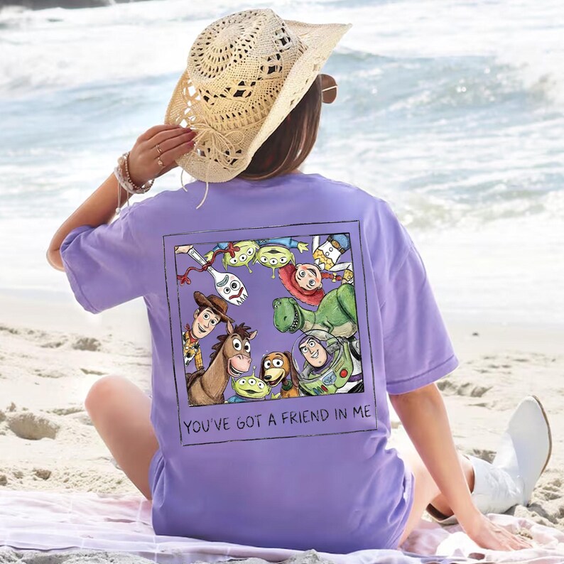 K&ouml;nnte beinhalten: Ein helllila T-Shirt mit einer farbenfrohen Grafik von Toy Story-Charakteren in einem quadratischen Rahmen mit dem Text "YOU'VE GOT A FRIEND IN ME". Ein Strohhut wird getragen. Das Bild wird am Strand aufgenommen.