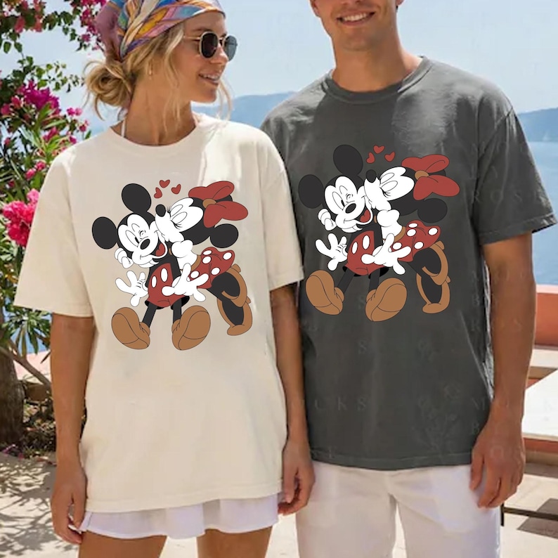 May include: Two t-shirts, one cream and one charcoal gray, each featuring a graphic of Mickey and Minnie Mouse embracing. Mickey is wearing brown shoes and Minnie has a red polka dot skirt and bow. Small red hearts float above them.