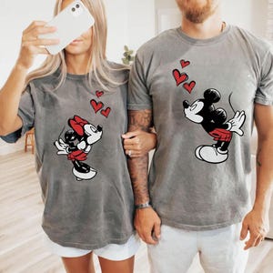 May include: Two grey t-shirts with Mickey and Minnie Mouse designs. Mickey is in black, red, and white, blowing kisses with red hearts above him. Minnie is in a red bow and skirt, also blowing kisses with red hearts.