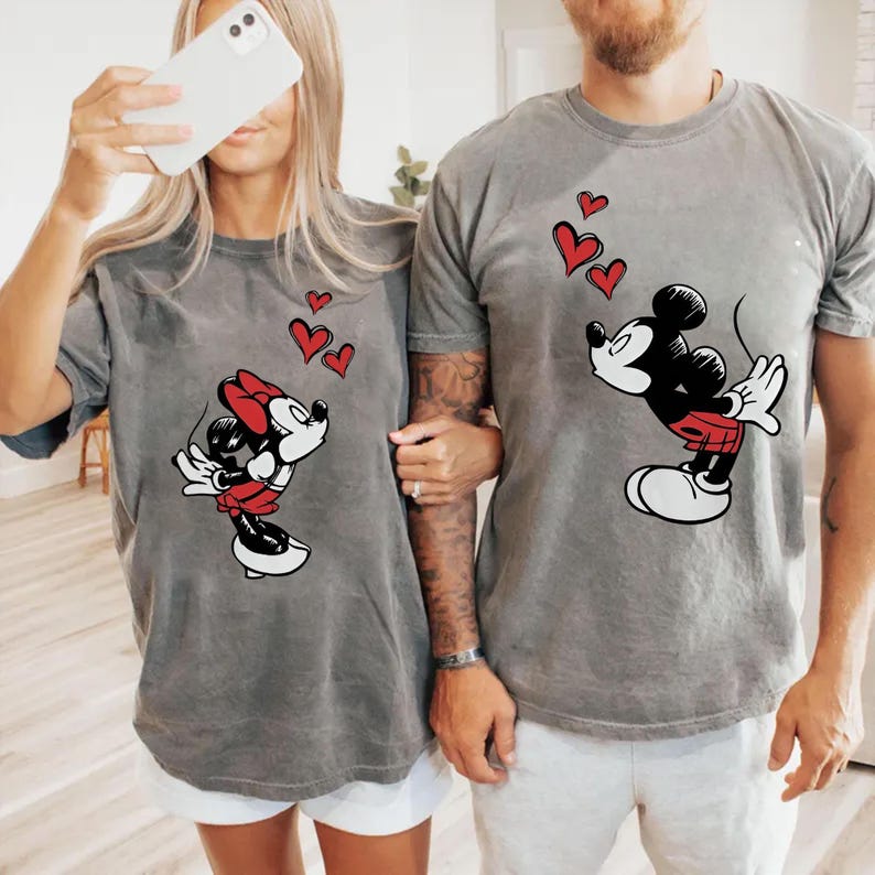 May include: Two grey t-shirts with Mickey and Minnie Mouse designs. Mickey is in black, red, and white, blowing kisses with red hearts above him. Minnie is in a red bow and skirt, also blowing kisses with red hearts.