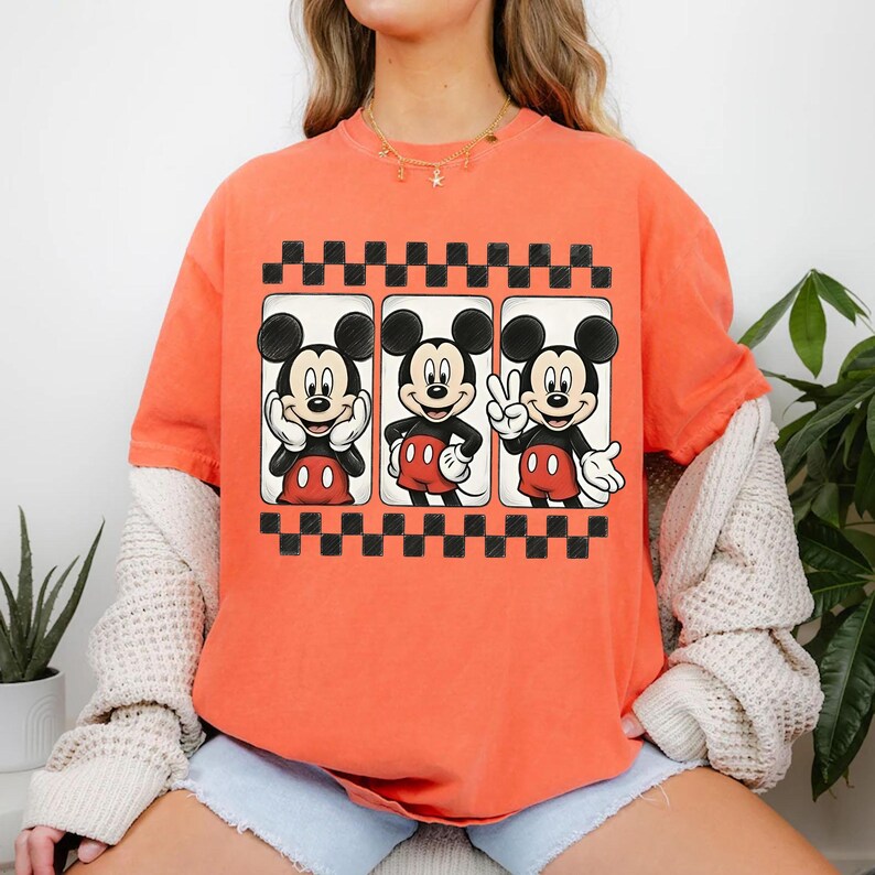 May include: Orange t-shirt featuring three Mickey Mouse images in black frames with a black and white checkered border. Mickey is depicted in various poses, wearing red shorts and white gloves.
