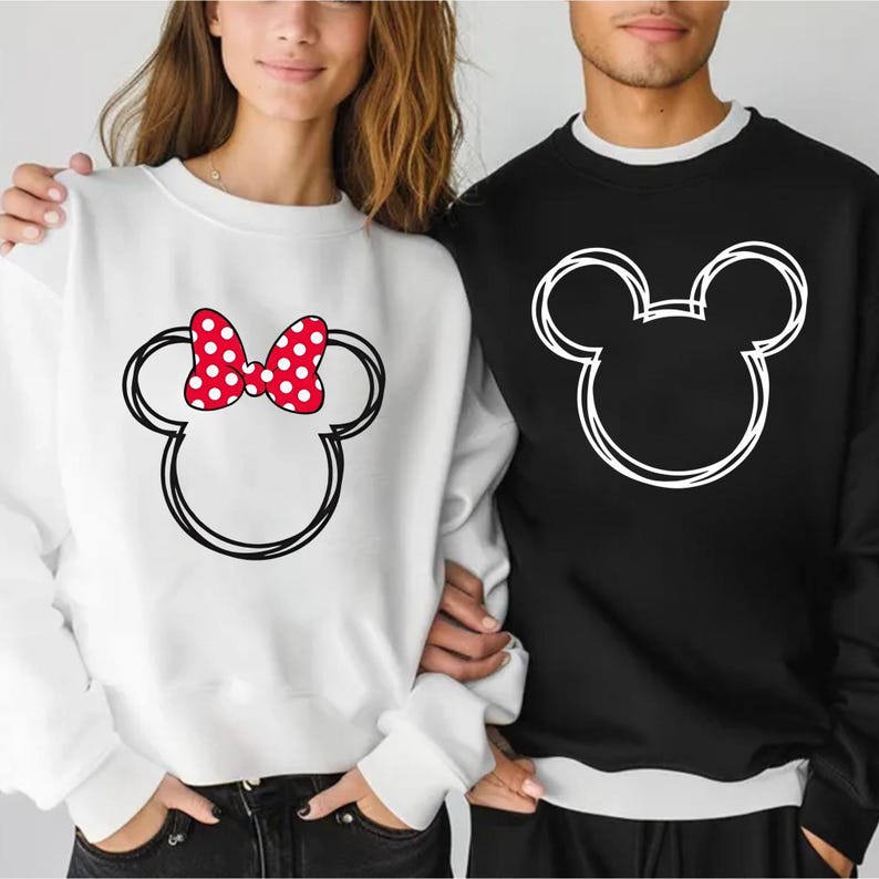 May include: A white sweatshirt featuring a black outline of a mouse head with a red polka-dot bow. Next to it, a black sweatshirt with a matching mouse head outline. The sweatshirts are part of a matching set.