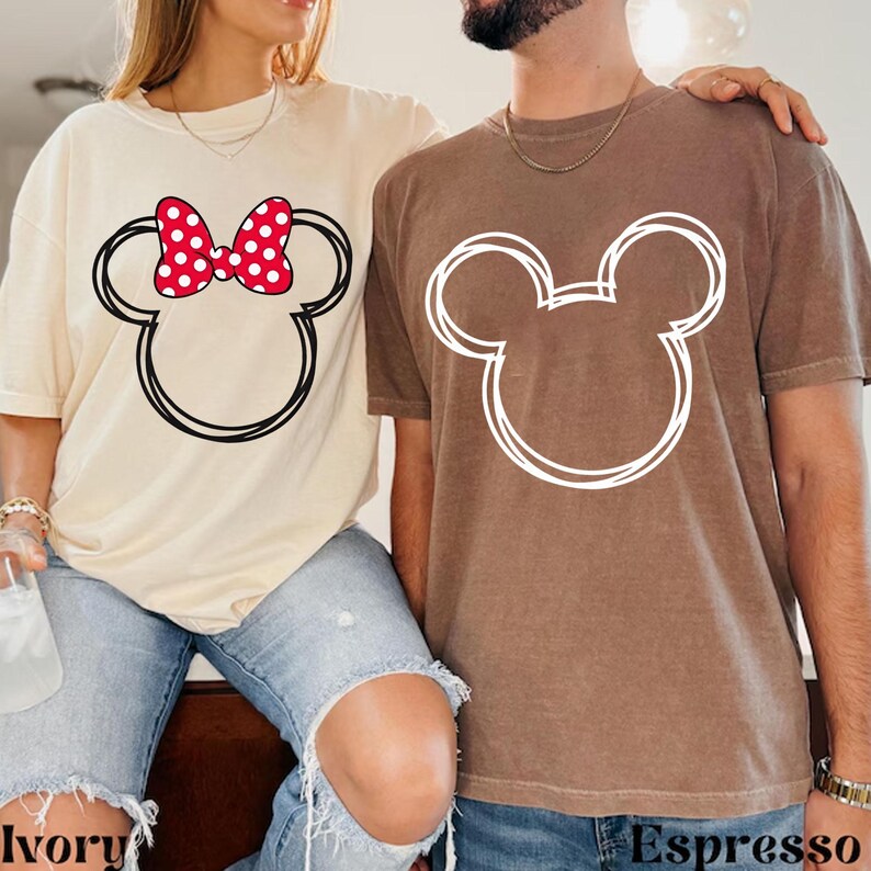 May include: Two t-shirts, one ivory and one espresso, featuring Mickey Mouse head outlines. The ivory shirt has a red polka dot bow. The espresso shirt has a white outline. The word "Ivory" is printed on the ivory shirt and "Espresso" on the espresso shirt.