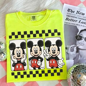 May include: Neon yellow t-shirt featuring three panels with Mickey Mouse illustrations. Each panel showcases a different pose of Mickey Mouse, with a black and white checkered border. The shirt has a crew neck and short sleeves.