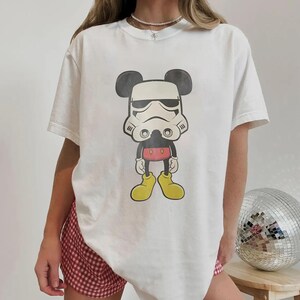 May include: White t-shirt with a cartoon design combining Mickey Mouse and a Stormtrooper helmet. The graphic features black ears, a white helmet, and yellow shoes. The shirt is a casual, short-sleeved style.