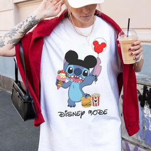 May include: White t-shirt featuring a cartoon Stitch character with a Mickey Mouse ear hat, holding an ice cream cone and a balloon. The shirt also has the words "Disney Mode" in black script.