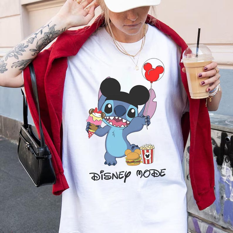 May include: White t-shirt featuring a cartoon Stitch character with a Mickey Mouse ear hat, holding an ice cream cone and a balloon. The shirt also has the words "Disney Mode" in black script.