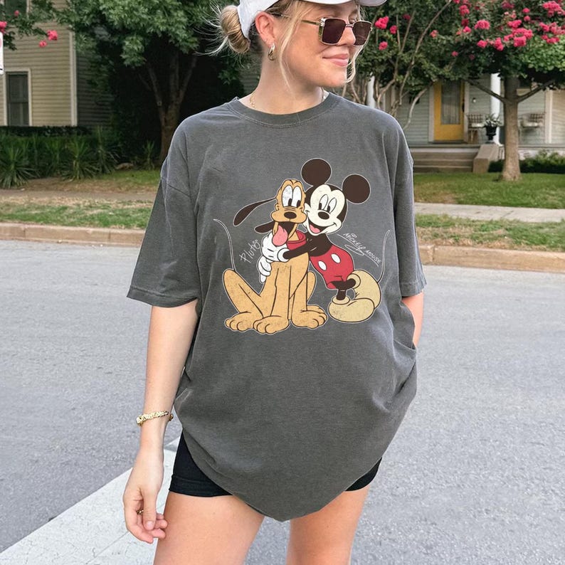 May include: Dark gray t-shirt featuring a graphic of Mickey Mouse hugging Pluto. Mickey Mouse is wearing red shorts and white gloves. Pluto is a light brown color. The shirt is a relaxed fit.