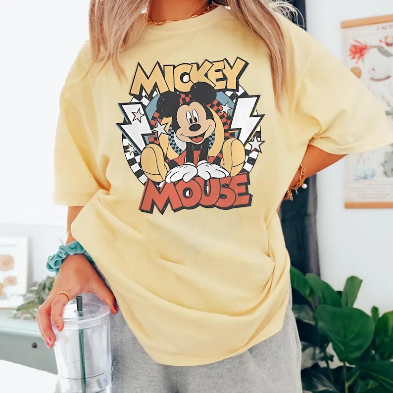 May include: Yellow t-shirt featuring a graphic of Mickey Mouse. The design includes the words "Mickey Mouse" in a retro font, with Mickey in the center. The shirt is a light yellow color.