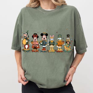 May include: Sage green t-shirt featuring a festive graphic of Disney characters dressed as toy soldiers. Goofy, Minnie Mouse, Mickey Mouse, Daisy Duck, Donald Duck, and Pluto are depicted on a skateboard, with holiday accents.