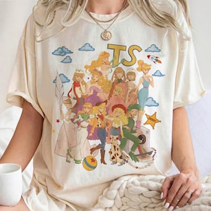 May include: A cream-colored short-sleeve t-shirt featuring a large graphic print of multiple animated female characters in various outfits. The design includes the letters TS, a rocket, a yellow star badge labeled SHERIFF, and a colorful ball.