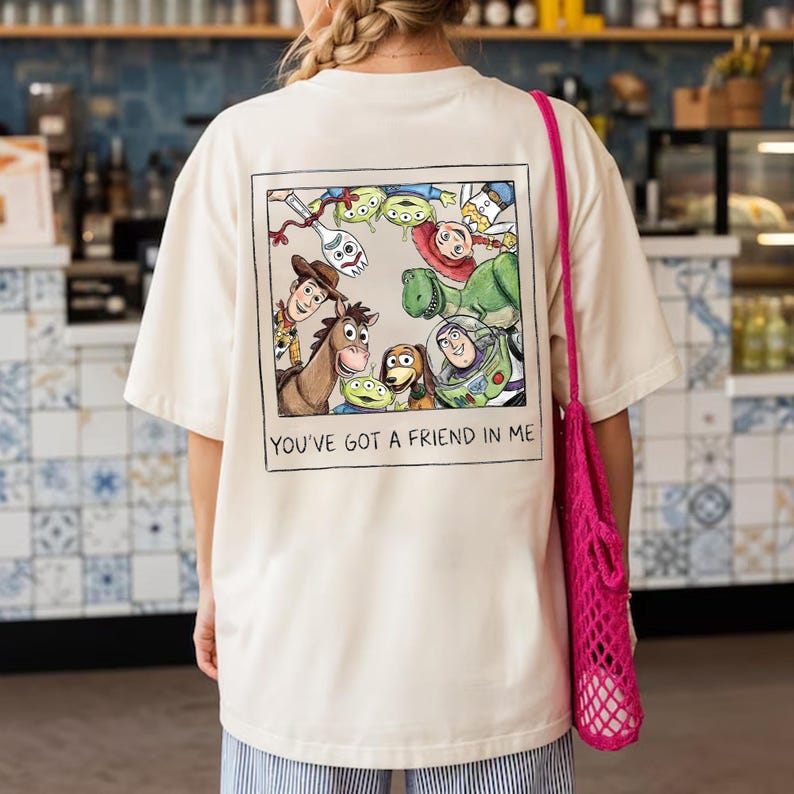 K&ouml;nnte beinhalten: Cremefarbenes T-Shirt mit einem gerahmten Cartoon-Motiv von Figuren aus dem Film "Toy Story". Die Grafik zeigt Woody, Buzz Lightyear und andere Charaktere. Der Text "YOU'VE GOT A FRIEND IN ME" steht darunter.