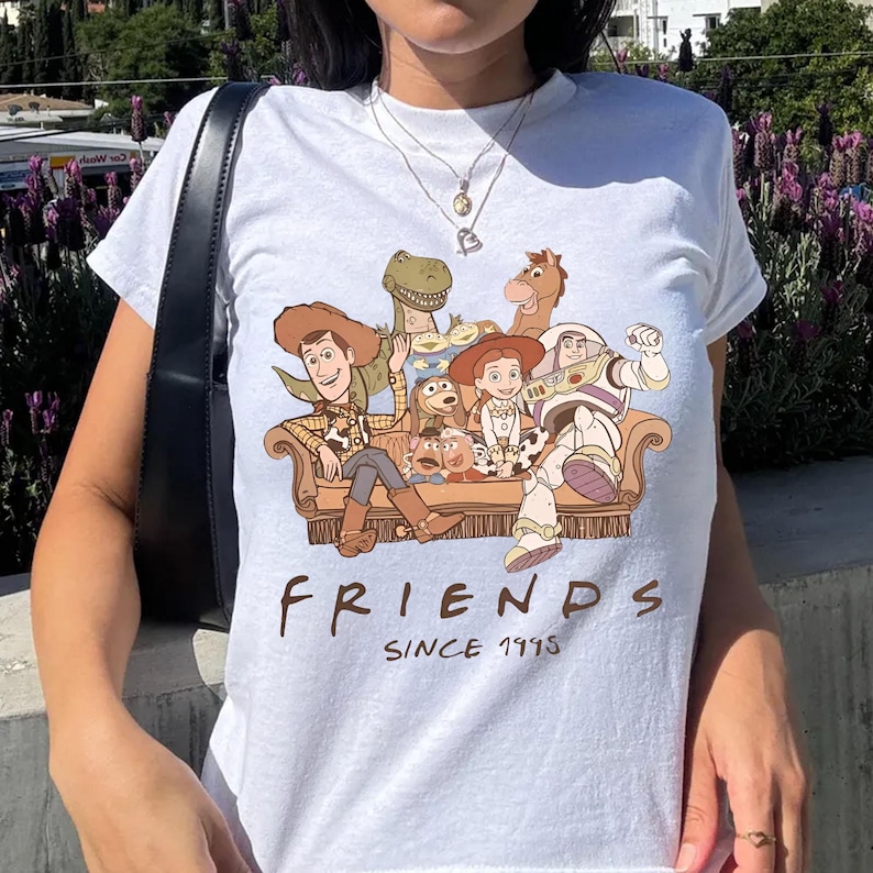 May include: White t-shirt featuring a colorful graphic of characters from the movie Toy Story, including Woody, Buzz Lightyear, and others, seated on a brown couch. The text "FRIENDS SINCE 1995" is printed below the graphic.
