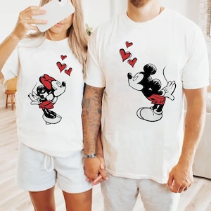 May include: Two white t-shirts with Mickey and Minnie Mouse designs. Mickey wears red shorts and Minnie a red bow. Both characters blow red heart shapes. The shirts are a matching set, ideal for couples.