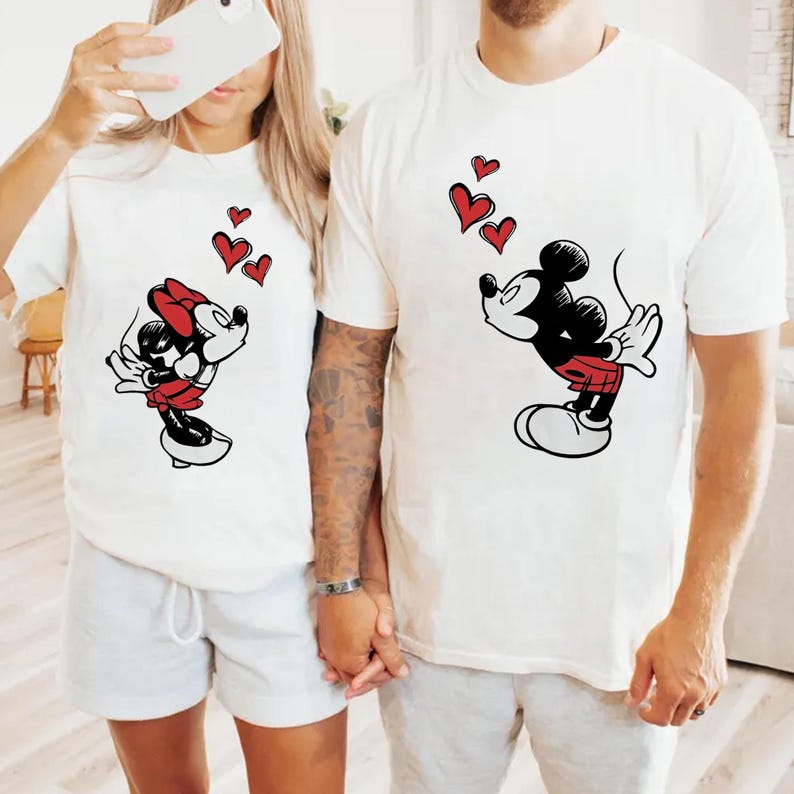 May include: Two white t-shirts with Mickey and Minnie Mouse designs. Mickey wears red shorts and Minnie a red bow. Both characters blow red heart shapes. The shirts are a matching set, ideal for couples.