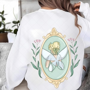 Könnte beinhalten: Weißes Sweatshirt mit einer Illustration einer Fee in einem goldgerahmten Spiegel. Die Fee hat blondes Haar und weiße Flügel, umgeben von grünen Blättern und rosa Herzen. Das Design befindet sich auf der Rückseite des Sweatshirts.