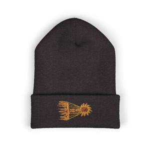 Könnte beinhalten: Dunkelgraue Mütze mit einem goldfarbenen, gestickten Design einer Sonne und eines Kamms. Die Mütze ist aus einem weichen, gestrickten Material gefertigt. Das gestickte Design befindet sich auf der Vorderseite des gefalteten Rands.