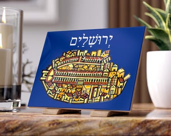 May include: A square, blue decorative tile featuring a stylized illustration of a city with yellow and brown buildings. The Hebrew word "Jerusalem" is written in white at the top. The tile is displayed on a wooden stand.