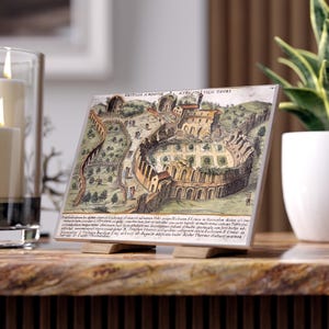 Può includere: Una piastrella in ceramica con una dettagliata illustrazione vintage di un anfiteatro e degli edifici circostanti. L'opera d'arte è nei toni del marrone, beige e verde, con testo in latino. La piastrella è esposta su un supporto di legno.