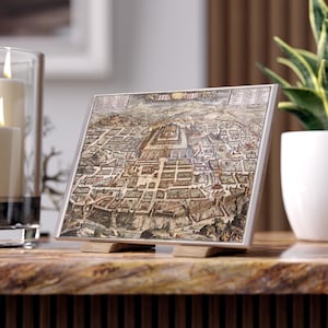 May include: A framed vintage map print depicting a detailed aerial view of a city, featuring buildings and streets in shades of brown, beige, and green. The print is displayed on a wooden stand, with a silver frame. The overall theme is historical cartography.