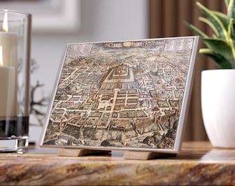 May include: A framed vintage map print depicting a detailed aerial view of a city, featuring buildings and streets in shades of brown, beige, and green. The print is displayed on a wooden stand, with a silver frame. The overall theme is historical cartography.