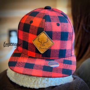 Könnte beinhalten: Rot-schwarz karierte Baseballkappe mit einem Lederaufnäher mit einem Hirschgeweih-Design und dem Text "Little Rocker". Die Kappe hat einen flachen Schirm und einen schwarzen Knopf oben. Der Markenname "Engraved Co" ist sichtbar.