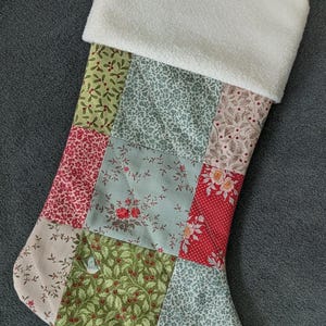 Peut inclure: Une chaussette de Noël en patchwork avec un revers en polaire blanc et une boucle de suspension. Le corps est fait de divers tissus à motifs rouges, verts et crème, avec des motifs floraux et botaniques.